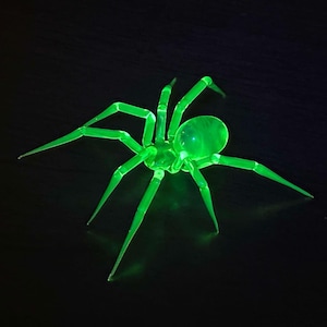 May include: A green glass spider sculpture with eight legs and a round body. The spider is glowing in the dark.
