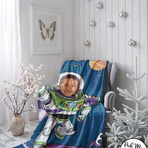 Puede incluir: Una manta personalizada de Buzz Lightyear con la cara de un ni&ntilde;o integrada en el traje del personaje. La manta presenta un fondo tem&aacute;tico espacial con planetas y estrellas, y el texto "YOUR NAME" en la parte inferior. La manta est&aacute; sobre un sill&oacute;n gris.