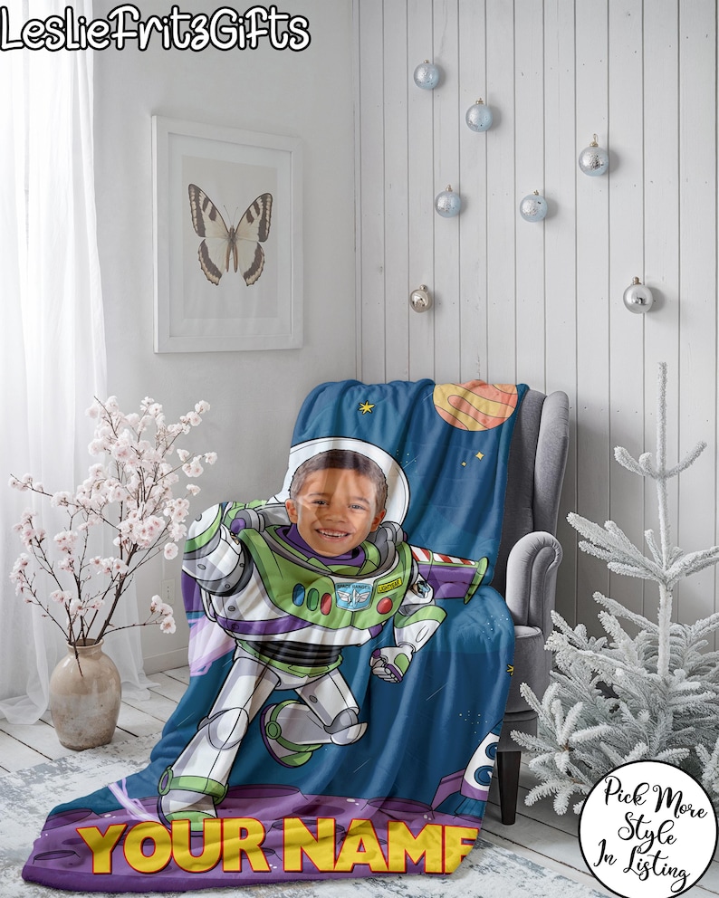 Puede incluir: Una manta personalizada de Buzz Lightyear con la cara de un ni&ntilde;o integrada en el traje del personaje. La manta presenta un fondo tem&aacute;tico espacial con planetas y estrellas, y el texto "YOUR NAME" en la parte inferior. La manta est&aacute; sobre un sill&oacute;n gris.