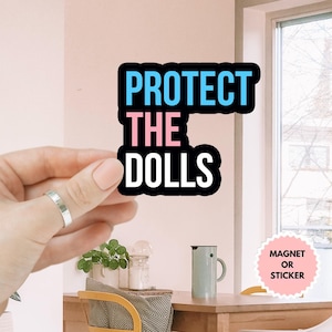 May include: A black and white sticker with the text "PROTECT THE DOLLS" in blue, pink, and white lettering. The sticker is being held by a hand. The sticker is available as a magnet or sticker.