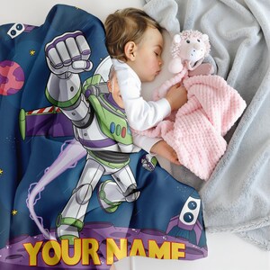 Puede incluir: Una manta azul con un dise&ntilde;o de dibujos animados de Buzz Lightyear y una escena espacial p&uacute;rpura y verde. El texto "YOUR NAME" est&aacute; en amarillo. Un ni&ntilde;o dormido est&aacute; en la manta, sosteniendo un juguete rosa y una manta rosa.