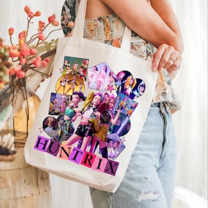 Może przedstawiać: Kremowa torba typu tote z kolażem kolorowych ilustracji w stylu anime przedstawiających grupę kobiet. Słowo "HUNTRIX" jest wydrukowane w stylu blokowym na dole torby. Torba ma dwa uchwyty.