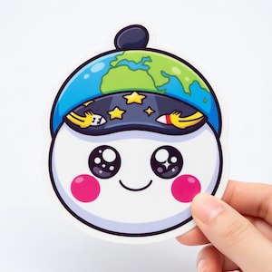 Puede incluir: Una pegatina troquelada que representa un personaje de dibujos animados con un cuerpo blanco, mejillas rosadas y ojos grandes y expresivos. El personaje lleva un sombrero azul con un diseño de globo terráqueo verde y azul y un borde de cielo estrellado. La pegatina está sujeta por una mano.