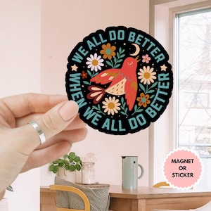 May include: A round black sticker with the text "WE ALL DO BETTER WHEN WE ALL DO BETTER" encircling a red bird with floral and celestial accents. The sticker is held by a hand.