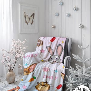 Puede incluir: Una manta azul claro y blanca con personajes de dibujos animados, estrellas y el nombre "EMILY". La manta est&aacute; sobre un sill&oacute;n gris. La imagen tambi&eacute;n incluye una impresi&oacute;n de mariposa enmarcada, adornos decorativos y el texto "LeslieFritzGifts".
