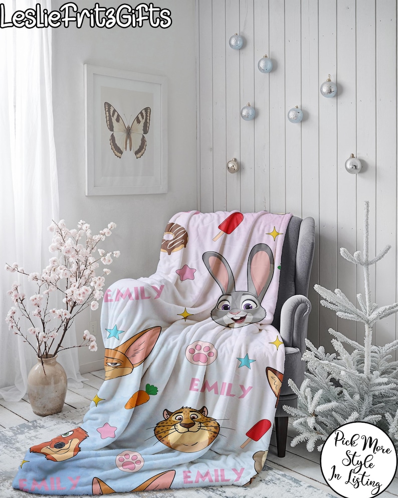 Puede incluir: Una manta azul claro y blanca con personajes de dibujos animados, estrellas y el nombre "EMILY". La manta est&aacute; sobre un sill&oacute;n gris. La imagen tambi&eacute;n incluye una impresi&oacute;n de mariposa enmarcada, adornos decorativos y el texto "LeslieFritzGifts".