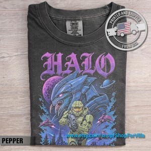Op de afbeelding: Een antracietgrijs t-shirt met een grafische print met het woord HALO in paarse letters. Het ontwerp bevat een groen gepantserde soldaat, een blauw buitenaards wezen, een paarse planeet en kleine ruimteschepen tegen een donkere sterrenhemel.