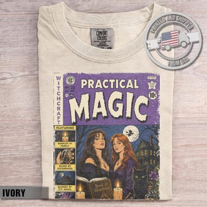 Könnte beinhalten: Ein elfenbeinfarbenes T-Shirt mit einer Vintage-Comic-Cover-Grafik mit dem Titel Practical Magic. Das Design zeigt zwei Frauen, eine schwarze Katze, ein Haus und ein Buch mit dem Titel Book of Shadows. Das T-Shirt hat ein Comfort Colors Etikett.