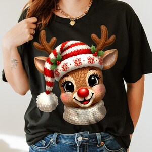 Puede incluir: Camiseta negra con un diseño de reno de dibujos animados. El reno tiene una nariz roja, un gorro a rayas rojas y blancas con copos de nieve y una bufanda blanca. El reno tiene cuernos marrones y está sonriendo.