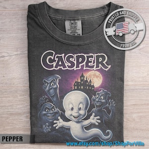 May include: A charcoal gray crew neck t-shirt featuring a graphic print of Casper the Friendly Ghost and three other ghosts in front of a haunted mansion under a full moon. The text CASPER is printed in a stylized white font above the characters.