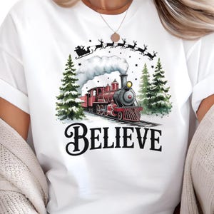 May include: White t-shirt featuring a festive design with a red and black train, evergreen trees, and Santa's sleigh pulled by reindeer. The word "BELIEVE" is printed in a bold, black font below the train.