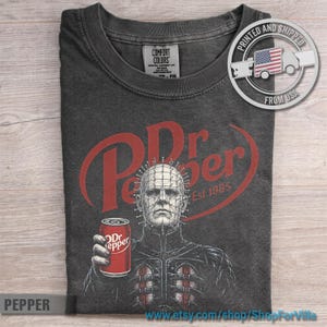 May include: A charcoal gray short-sleeve t-shirt featuring a graphic of the character Pinhead holding a red Dr Pepper soda can. The design includes the red Dr Pepper logo and the text Est. 1885. The shirt has a Comfort Colors brand neck label.