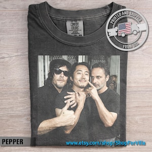 May include: A charcoal gray crew neck t-shirt featuring a black and white photographic print of three individuals. The shirt has a Comfort Colors brand label at the inner neckline and a circular graphic in the top right corner with a flag icon.