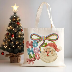 Puede incluir: Un bolso tote de color crema con un diseño festivo que incluye las palabras "HO HO HO", una cara de Papá Noel y un lazo. A la izquierda hay un pequeño árbol de Navidad con adornos rojos y una estrella.