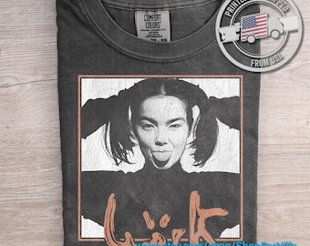 May include: A charcoal gray crew neck t-shirt featuring a black and white graphic print of a person with pigtails sticking their tongue out. Below the portrait is stylized text. The shirt has a Comfort Colors brand label at the inner neckline.