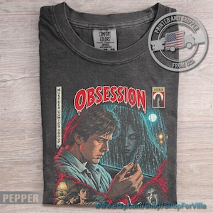 May include: A charcoal gray t-shirt featuring a graphic print styled like a comic book cover. The text reads "OBSESSION" in large red letters, with "MUSTEREE COMICS GROUP" on the side and a "HORROR" box. The illustration depicts a person holding a small object.