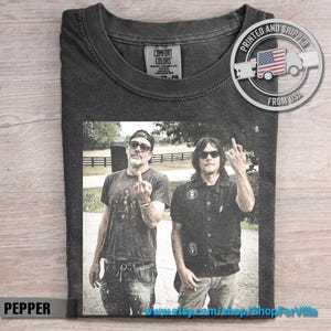 May include: A charcoal gray t-shirt featuring a graphic print of two individuals wearing sunglasses and giving the middle finger. The shirt has a crew neckline and a Comfort Colors brand label on the inside collar, with a circular logo in the corner.