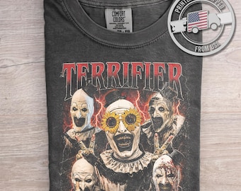 Puede incluir: Una camiseta de cuello redondo gris carbón con un estampado gráfico de Art the Clown de la serie de películas Terrifier. El diseño incluye la palabra Terrifier en letras rojas sobre cinco figuras de payasos, uno con gafas de girasol, sobre un fondo de rayos rojos.