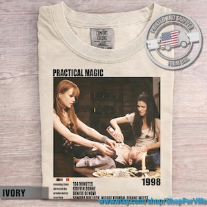 Puede incluir: Una camiseta de color marfil con un estampado gráfico de la película Practical Magic de 1998. El diseño incluye una escena con dos personas de pie sobre alguien acostado, el título de la película, detalles de producción y el año 1998 en negro.