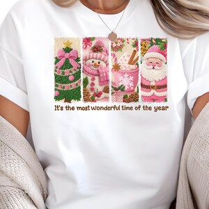 Puede incluir: Camiseta blanca con un diseño navideño. El diseño incluye un árbol de Navidad, un muñeco de nieve, chocolate caliente y Papá Noel, con detalles en rosa y verde. El texto dice "It's the most wonderful time of the year."