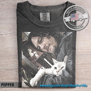 May include: A charcoal gray short-sleeve t-shirt featuring a graphic print of a person with dark hair holding a white cat. The shirt has a crew neckline and a Comfort Colors brand label on the inside collar. The graphic is centered on the chest.