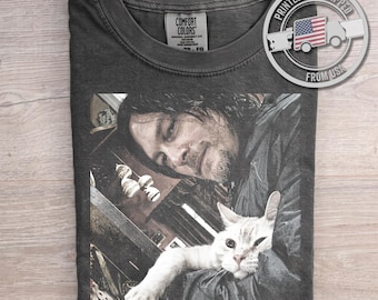 May include: A charcoal gray short-sleeve t-shirt featuring a graphic print of a person with dark hair holding a white cat. The shirt has a crew neckline and a Comfort Colors brand label on the inside collar. The graphic is centered on the chest.