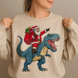 Könnte beinhalten: Beiges Sweatshirt mit einem festlichen Motiv. Es zeigt den Weihnachtsmann in rotem Anzug und Hut, der einen blauen Tyrannosaurus Rex reitet. Der Weihnachtsmann zeigt mit der rechten Hand und trägt einen Sack mit Geschenken. Das Maul des Dinosauriers ist geöffnet und zeigt seine Zähne.