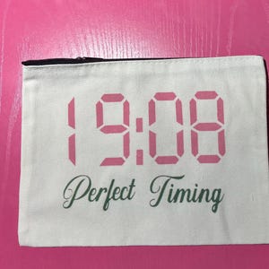 May include: A white canvas pouch with a black zipper, featuring the pink digital display of 19:08 and the words "Perfect Timing" in green script. The pouch is rectangular and sits on a pink surface.