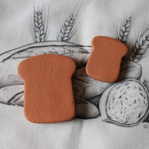 May include: Two terracotta-colored, bread-shaped objects are displayed on a fabric surface with a wheat and bread illustration. The objects are a light brown color and have a textured surface. The background features a black and white drawing of wheat and bread.