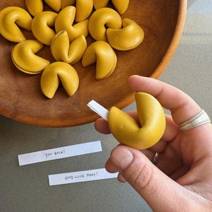May include: A close-up of a hand holding a yellow fortune cookie. The cookie is open and the paper inside is visible. The paper says "You rock!" and "Good luck today!" There are other fortune cookies in a wooden bowl in the background.