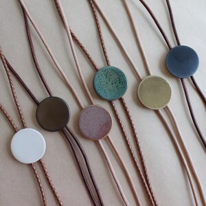 May include: A collection of bolo ties with circular ceramic pendants in various colors, including white, brown, green, and blue. The ties feature braided and smooth leather cords, offering a rustic and handcrafted aesthetic.