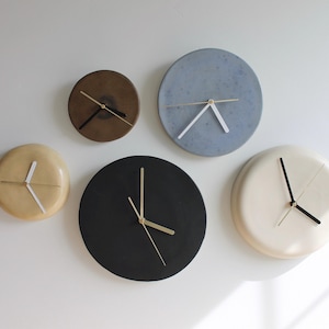 May include: A collection of five minimalist wall clocks in various colors and sizes. The clocks have round faces in shades of brown, blue, black, and cream. Each clock features simple hands in contrasting colors, such as gold and white.