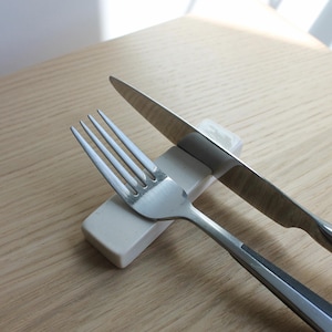 May include: A stainless steel fork and knife rest on a small, rectangular, off-white ceramic holder. The knife is angled across the holder, and the fork is positioned below it. The background is a light-colored wooden surface.