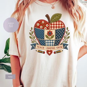 Peut inclure: Un t-shirt ivoire avec un grand motif de pomme en patchwork. Le design de la pomme comprend des motifs floraux, à carreaux et à pois en rouge, bleu et blanc. Sous la pomme se trouve une bannière avec le texte TEACH INSPIRE GROW, entourée de deux crayons et d'un petit cœur.