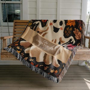 May include: A woven throw blanket featuring a large cream-coloured ghost graphic against a black background with orange, brown, and red autumn leaves. A central tan banner displays the text The Davis Family Est. 2025. The blanket has fringed edges.