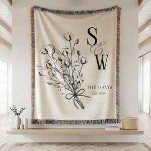 May include: A cream-colored woven throw blanket with a fringed edge featuring a black line-art illustration of a cotton bouquet, the letters S and W, the text THE DAVIS, and the text Est. 2026 printed in a serif font on the right side.