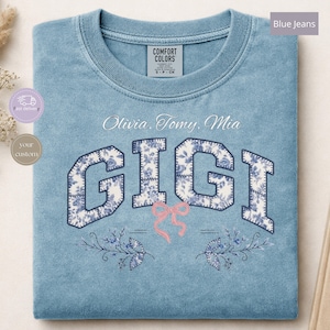 May include: A blue crewneck sweatshirt featuring the name GIGI in large block letters with a white and blue floral pattern. Above the name are the words Olivia, Tomy, Mia. Below the name is a small pink bow and two blue floral sprigs.