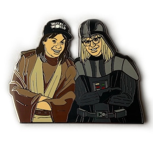May include: A black and white enamel pin featuring two characters. The character on the left is wearing a brown robe and a black hat with the words "Wayne's World" on it. The character on the right is wearing a black cape and a black helmet with a white face mask. The character on the right has a white beard and is wearing glasses.