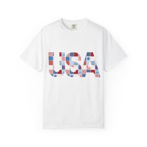 May include: A white short-sleeve t-shirt featuring the letters USA printed in a patchwork design. The letters are composed of various red, white, and blue patterns, including stars, stripes, and checkered squares, creating a patriotic aesthetic.