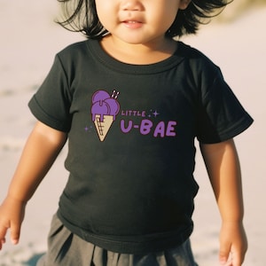 May include: A child wearing a black t-shirt with a purple ice cream cone graphic and the text "Little U-Bae".