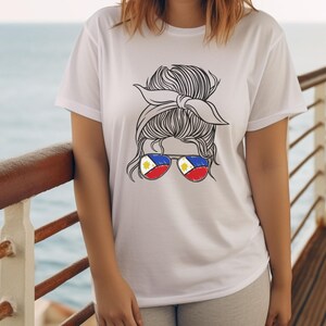 May include: White t-shirt featuring a graphic of a woman's hair styled in a bun with a headband. The sunglasses have the colors of the Philippine flag. The background shows a body of water and a wooden deck.
