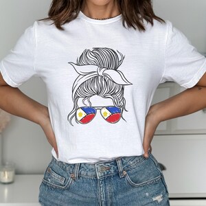 May include: White t-shirt featuring a black and white graphic of a woman's updo hairstyle with sunglasses. The sunglasses have the colors of the Philippine flag. The shirt is a crew neck and is made of a soft material.