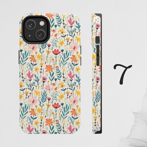 May include: A white phone case with a floral pattern featuring pink, yellow, orange, and blue flowers. The case has a black camera cutout and a black border.