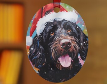 May include: Oval Christmas decoration featuring a black dog wearing a Santa hat. The dog has brown eyes and a pink tongue. The decoration is suspended from a red ribbon against a blurred background of a Christmas tree and a bookshelf.