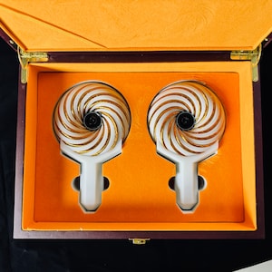 Puede incluir: Dos objetos decorativos de color blanco y dorado con un diseño de espiral en una caja de madera marrón con un forro de terciopelo naranja.