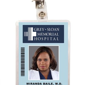 May include: A blue and white plastic ID badge with a photo of a woman with dark hair and a white lab coat. The badge reads "GREY + SLOAN MEMORIAL HOSPITAL" and "MIRANDA BAILE, M.D. Chief of Surgery 7002-2901".