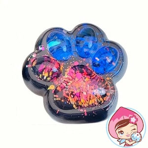 May include: A paw-shaped resin item with a black border. The paw pads are filled with colorful glitter and blue resin. The item is on a white background.