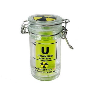 Op de afbeelding: Een glazen pot met een geel en zwart radioactief symbool op de deksel. De pot bevat een gele vloeistof en een etiket met de tekst "U Uranium (238-028) Radioactief III Activiteit: 12,358 Bq".
