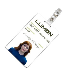 May include: A white plastic ID card with the text "LUMON" in green letters. The card has the text "hello, my name is HELLY RIGGS SVR'D ACCESS" and "employee code 08-988". A photo of a woman with red hair is in the top left corner of the card. The text "signature" is below the photo and a handwritten signature is below that.