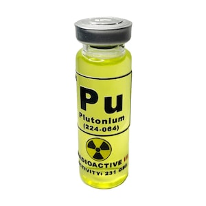 May include: A small glass vial with a yellow label that reads "Pu Plutonium (224-064)" and a black and yellow radioactive symbol. The vial contains a clear yellow liquid.